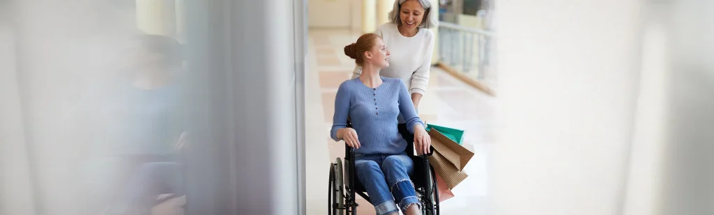 Pflegerin schiebt junge Frau im Rollsltuhl mit Einkaufstaschen vor Haus