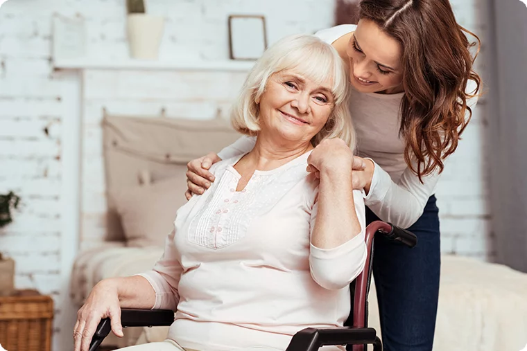 Frau im Rollstuhl lacht und wird von jüngerer Frau umarmt